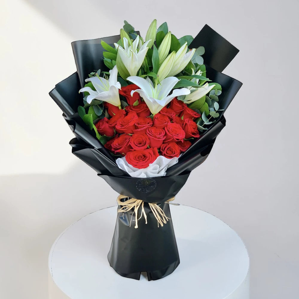 Bouquet of red roses and white lilies wrapped in black paper on a white stand.